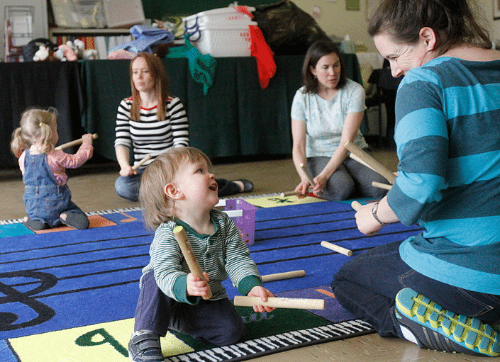 A toddler sitting on the floor in front of a teacher playing with two drum sticks