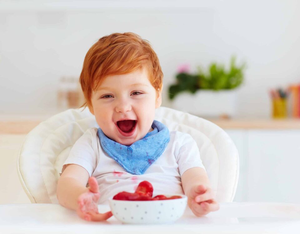 Happy red-headed child (toddler) in a high chair eating strawberries. Babysitting