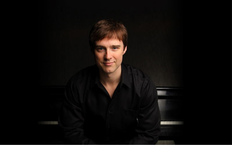 Michael Cavanaugh wearing all balck, sits in front of a grand piano