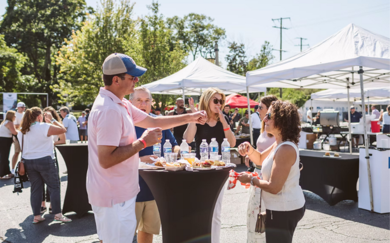 Five people eating and drinking at a high boy table with a festival setting in the background.