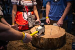 3 people standing around a wood stump with nails in it. One person is hitting a nail into the stump, a woman wearing a german style dress is waiting for her turn while a third person is holding a beer