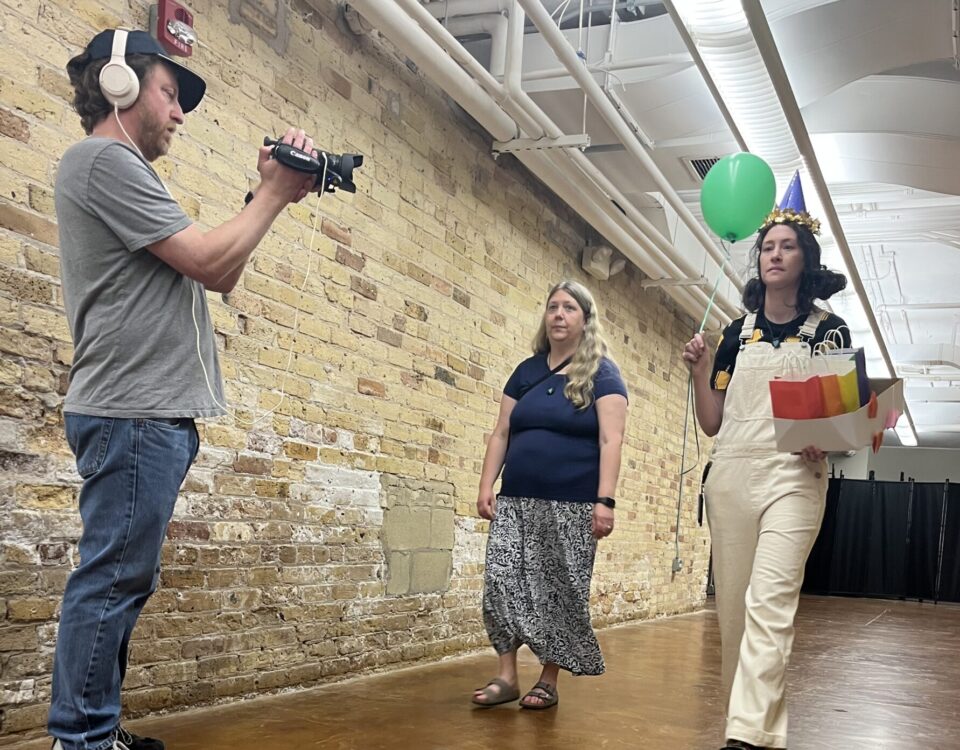 Jeremy Kruse holding a camera videotaping a girl walking down a hallway wearing a birthday hat and holding a balloon and gift bags
