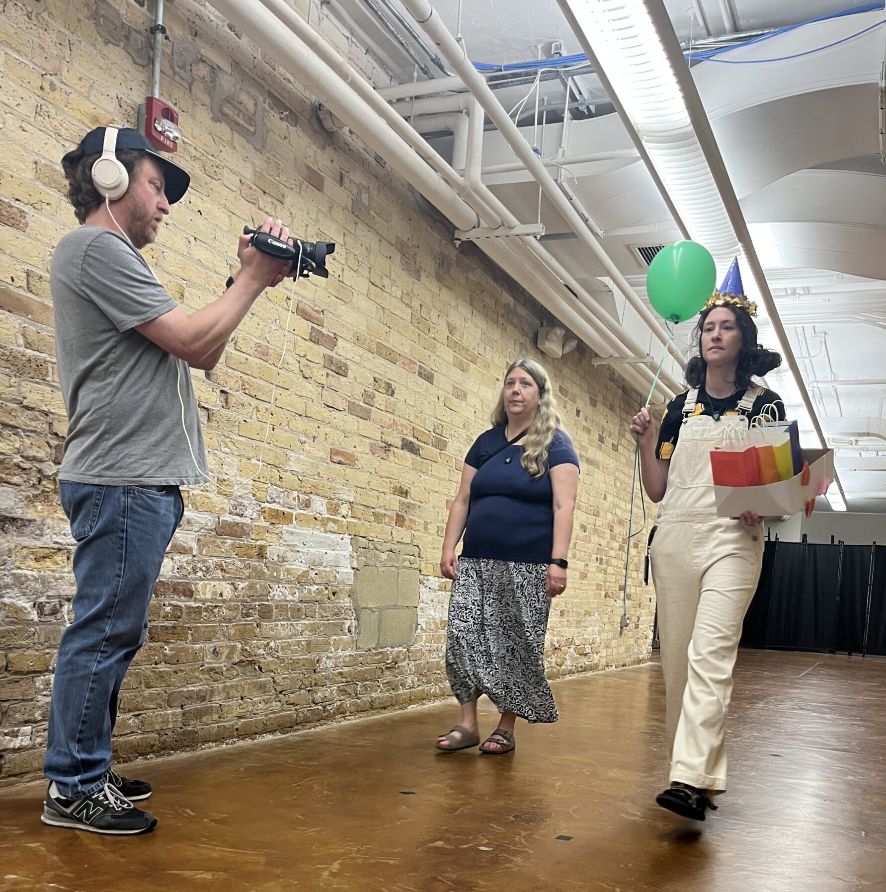 Jeremy Kruse holding a camera videotaping a girl walking down a hallway wearing a birthday hat and holding a balloon and gift bags