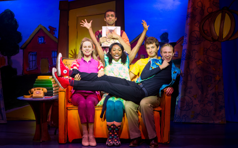 The cast of Pete the cat is on stage. A group of 5 boys and girls are sitting on a colorful yellow couch. One boy is standing behind the couch holding a photo of a cat. The girl sitting in the middle of the couch has her hands up in the air and an excited look on her face. Everyone is wearing colorful clothes, smiling and looking directly at the camera.