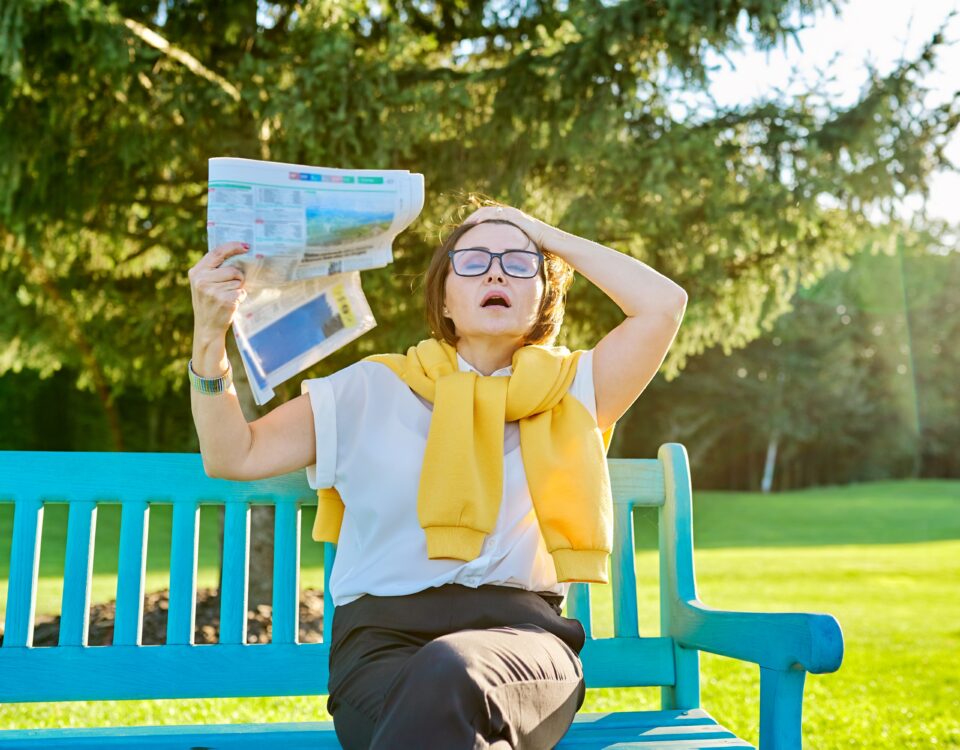 WOman on park bench waves herself with a newspaper while having a hotflash