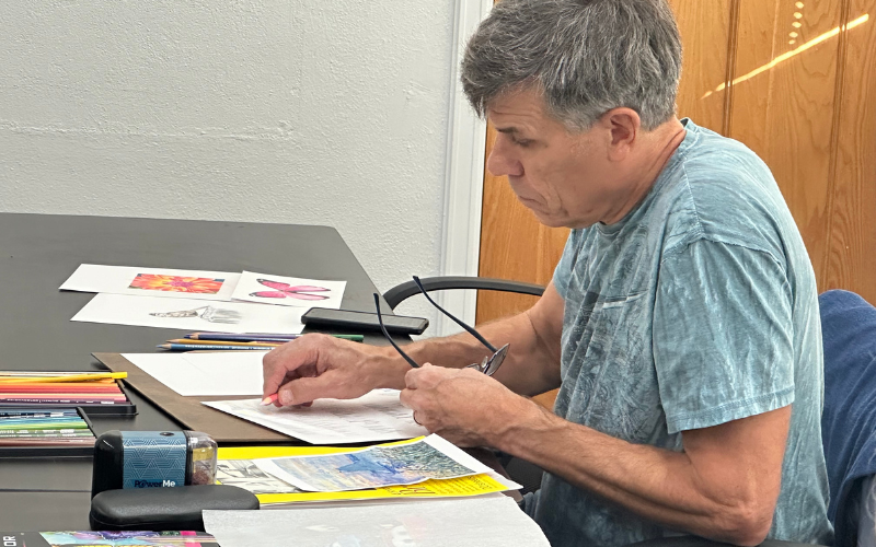 A man in the Color Pencil Art class at Gorton. The photo is a view from the side- he is holding a pencil and working on a sketch. On the able around him are different color pencils and some completed drawings.