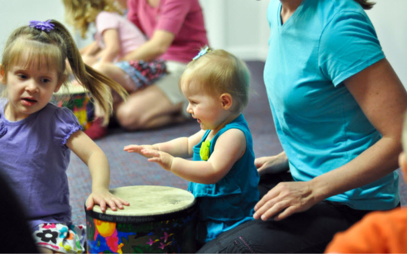 Small children sitting on the floor with a hand drum and adults sitting around them