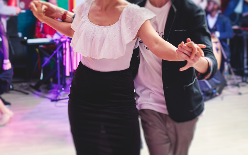 An action shot of a man standing behind a woman who is facing the camera- they have their arms out to the sides and their hands are clasped - they are appear to me moving towards the camera as they dance