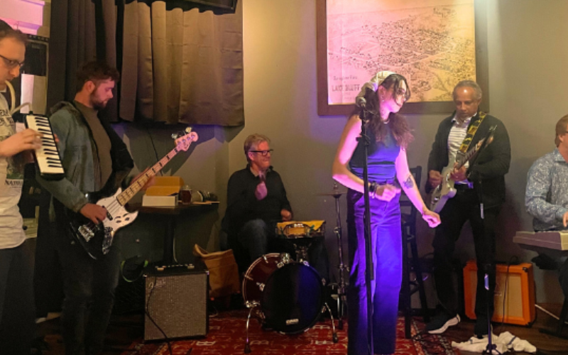 A group of six people in a room with musical instruments; a man with a keytar, two men with guitars, one man on drums, a woman standing in the middle with a micrphone and a man on keyboards to the right of her