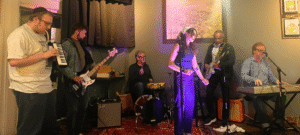 An image of five people playing music together; one man playing keytar, two men playing guitar, one man playing the keyboard and a woman standing in the middle with a microphone