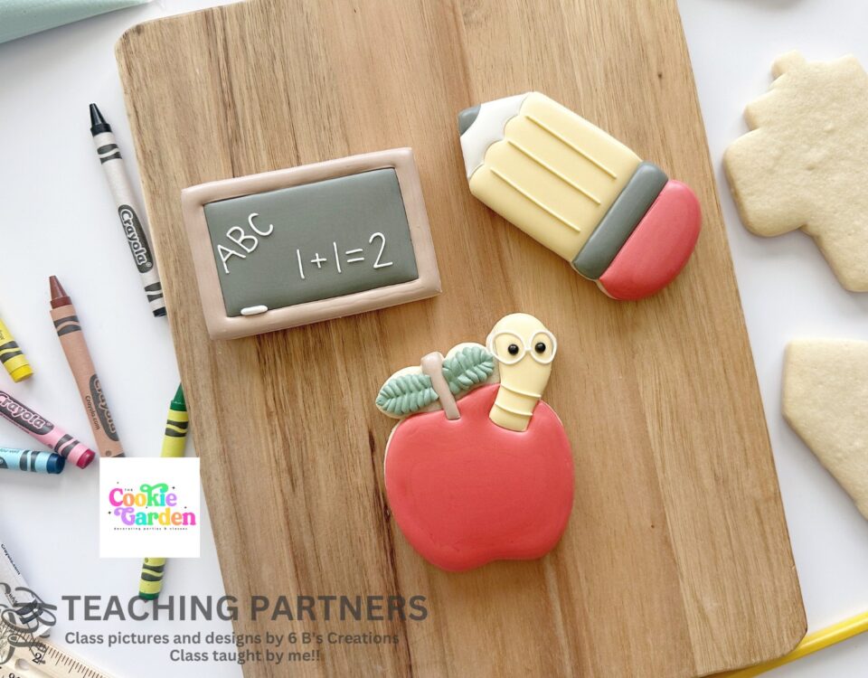 3 cookies on a wooden cutting board decorated to look like a small chalk board, an apple with a cute little worm sticking out and a pencil. There are plain sugar cookies and crayons on either side of the cutting board.