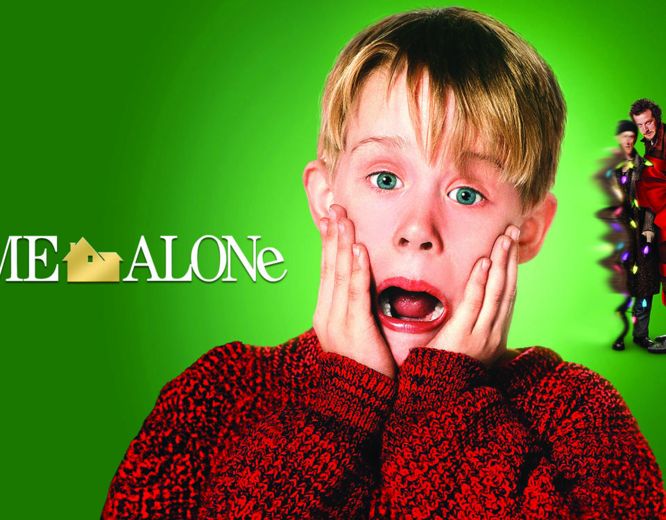 Promotional photo for Home Alone: A young boy wearing a red sweater, has his mouth open wide and his hands on either side of his face as if he's screaming in fear and is looking right at the camera, Behind him to his right are two sinister looking men wrapped in christmas lights and bows