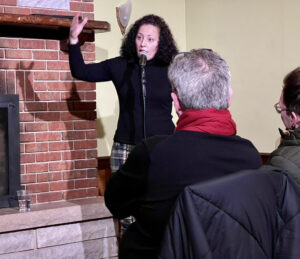 unnamed-1-2-300x259-1 Woman in a black turtleneck talking into a microphone in front of a small audience at a storytelling event