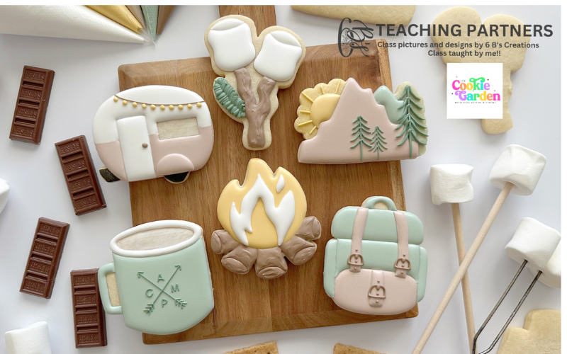 Cookies sitting on a cutting board that have been decorated to look like a campfire, a small camper, a mug, marshmallows on a stick and the sun setting above the trees. The cookies are surrounded by pieces of chocolate and marshmallows on sticks.