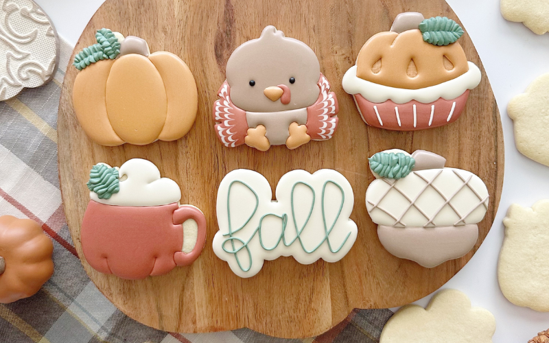 Six cookies very cutely decorated and sitting on a cutting board. They are decorated to look like a pumpkin, an owl, an acorn, a mug and the word 'fall'. There are plain sugar cookies on the side of the cutting board and a plaid tablecoth underneath.