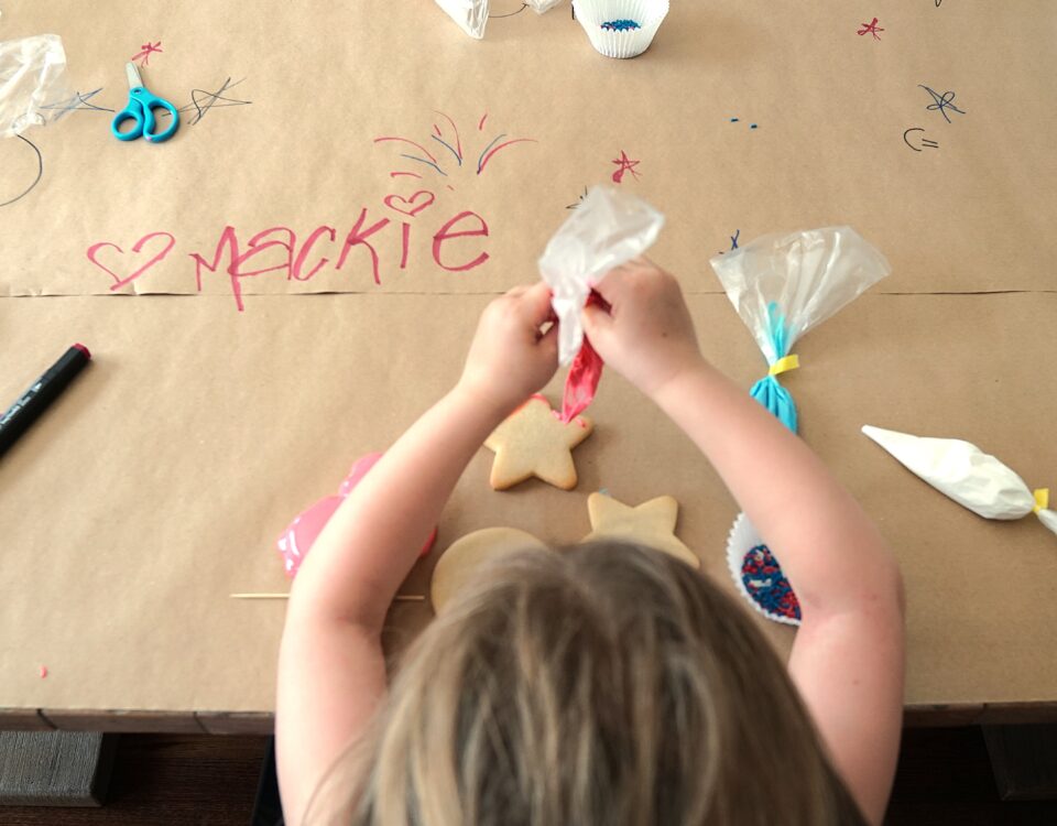 A photo from above a young child's head- they are seated at a table covered in paper with writing on it and are piping frosting onto a plain sugar cookie