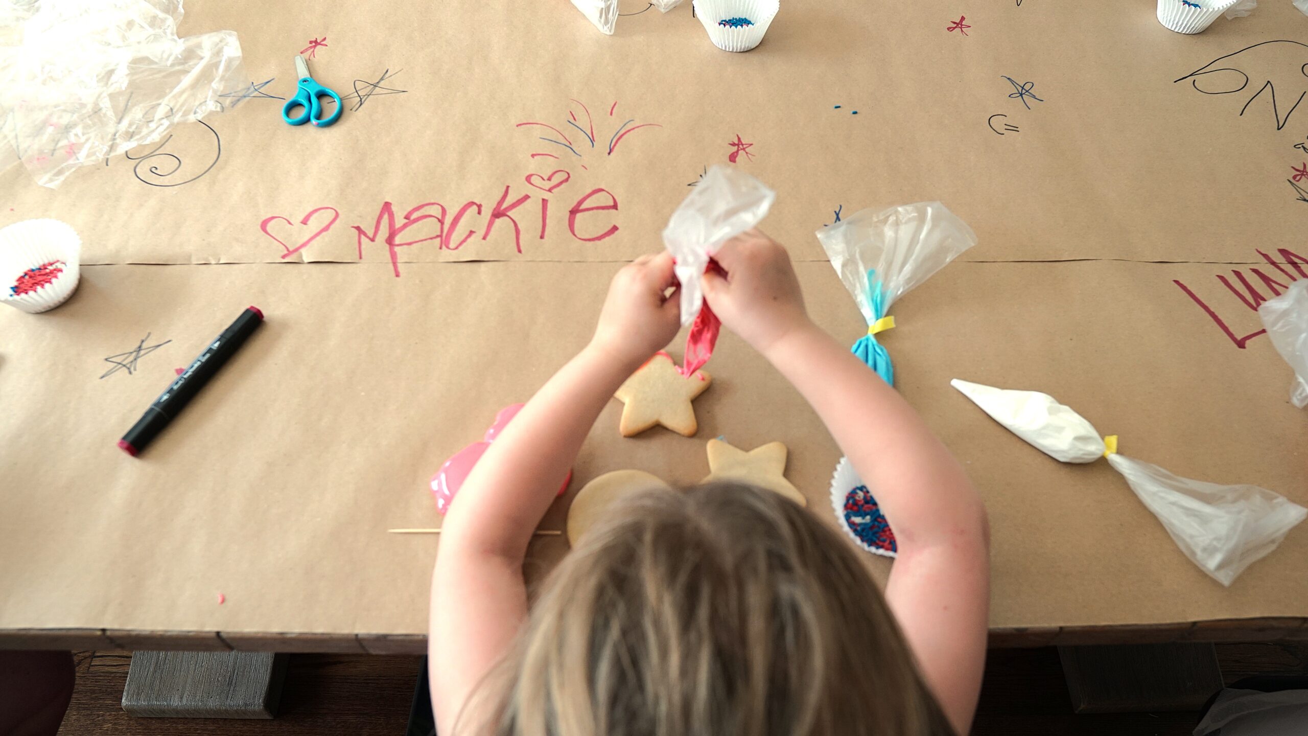 A photo from above a young child's head- they are seated at a table covered in paper with writing on it and are piping frosting onto a plain sugar cookie