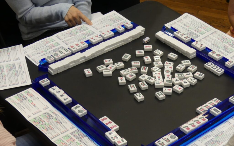 A view of the top of a black card table with 4 sets of mahjong tiles and scorecards. A person sitting at the table is holding their mahjong scorecard and appears to be reading information off of it.