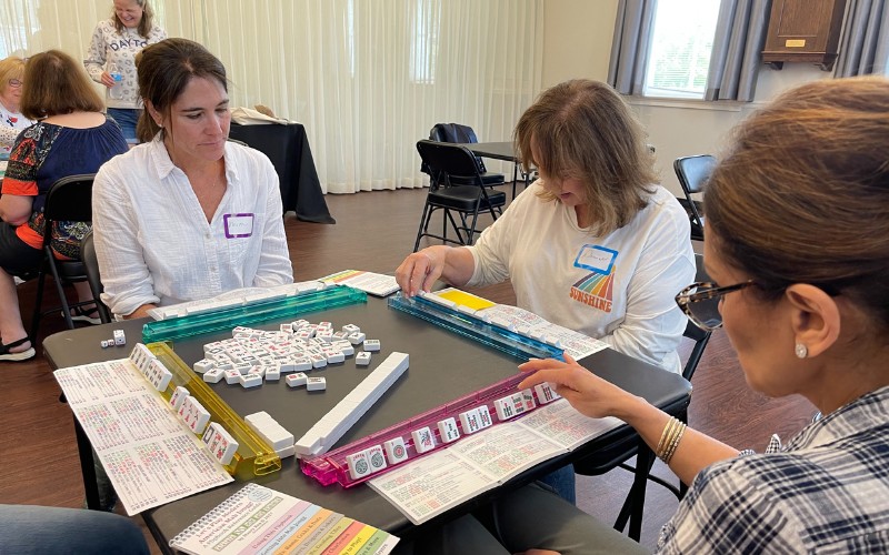 3 women sitting at a table playing mahjong- each has a set of tiles in front of them as well as a scorecard