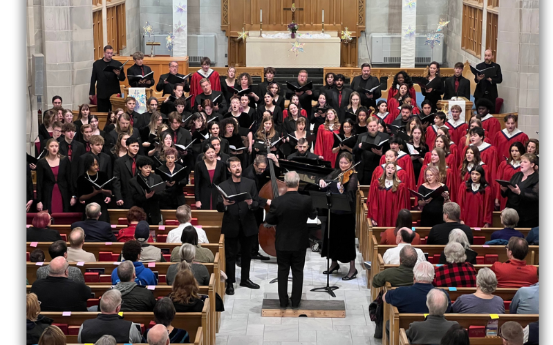 Members of the St. Charles are standing in a church and singing. They are all dressed in red or black. Gathered at the front of the group are musicians playing instruments and a conductor stands at the very front with his arms up