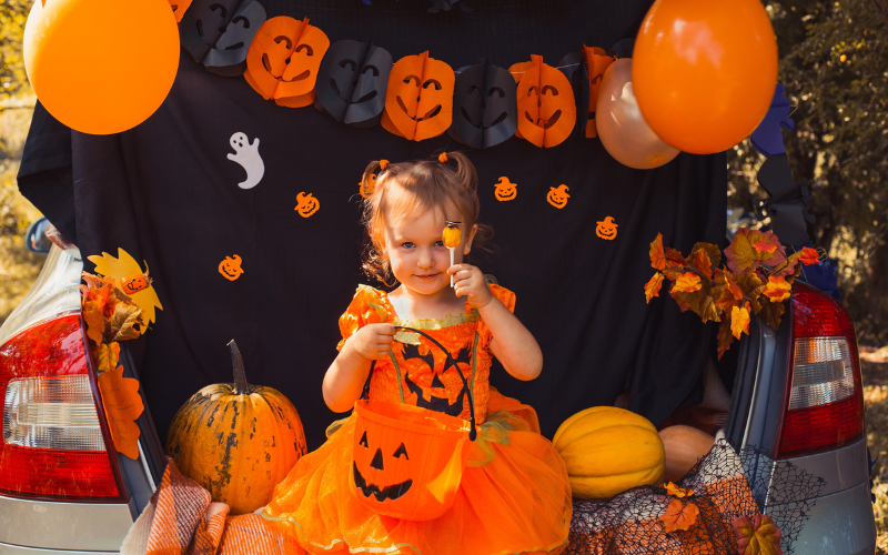 A toddler aged girl dressed in a jack o' lantern dress posing in front of her trunk which has black background, a streamer of cutout jackout lanterns and orange balloons. She is holding up her treat, a lollipop for the camera to see.