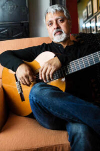 fareed haque seated on a cushioned orange chair holding the his guitar and looking at the camera