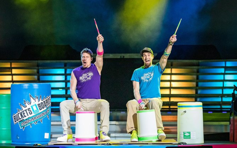 Both members of Buckets N Boards performing onstage. They are each sitting on top of a bucket holding up one drumstick in their left hands. The are other buckets on the stage and colorful lights are in the background.