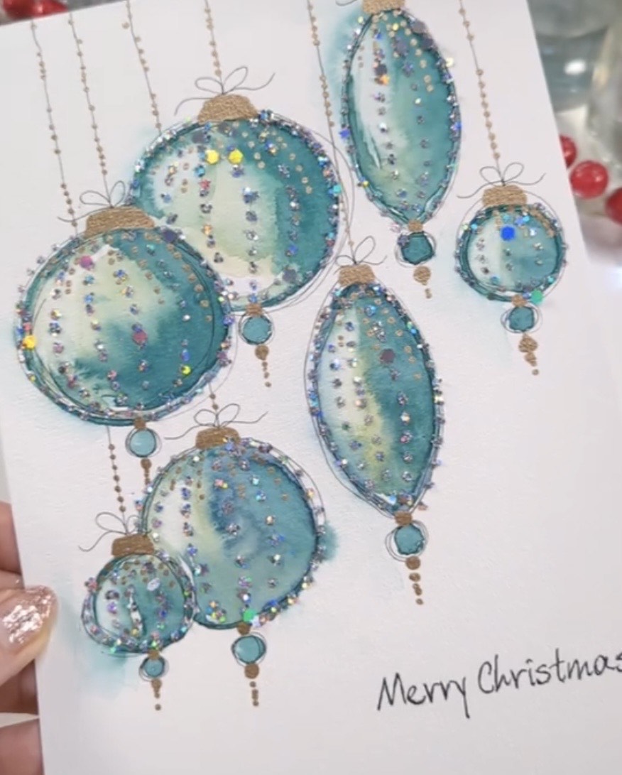 Handmade holiday card featuring painted ornaments on the front that are decorated with small jewels and made to look like they are hanging. at the very bottom the card reads 'Merry Christmas'