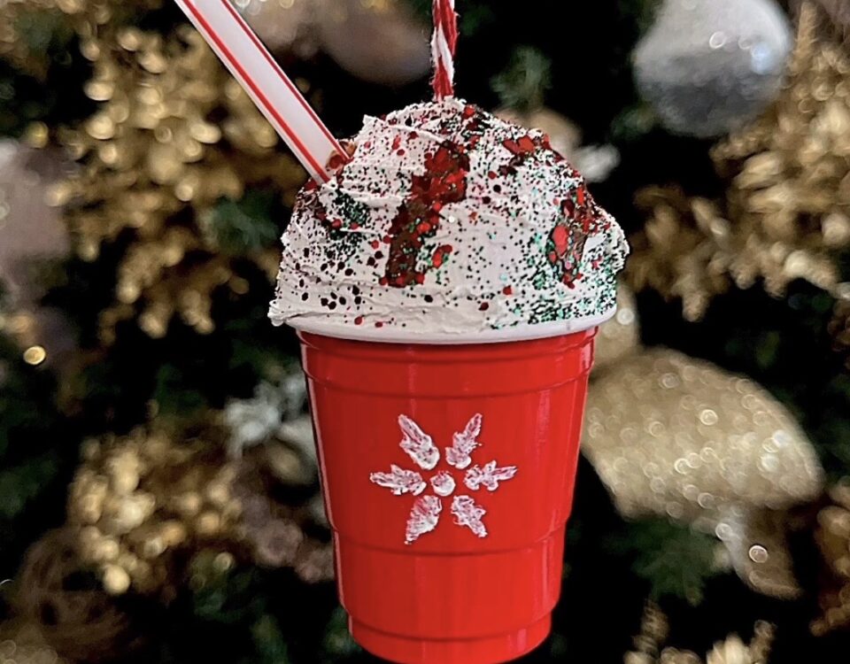 An image of a DIY holiday ornament hanging from a tree- it looks like a small cup of hot chocolate with a big pile of whip cream on top., The cup is red with a snowflake on it.