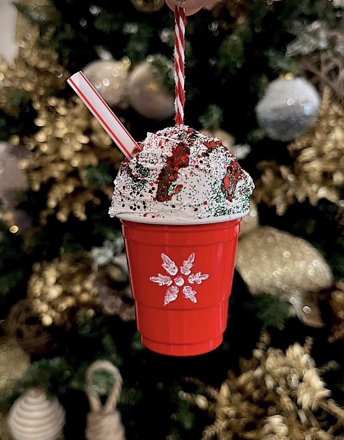 An image of a DIY holiday ornament hanging from a tree- it looks like a small cup of hot chocolate with a big pile of whip cream on top., The cup is red with a snowflake on it.