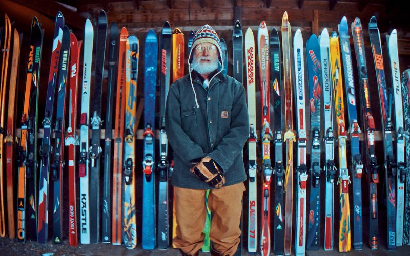 Older Man in a coat and colorful ski hat standing in front of a wall of skis