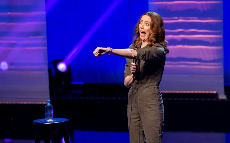 Comedian Leah Rudick is on stage in her 'spiraling' stand up special. Her mouth is open wide and she has her left arm extended out towards the audience.