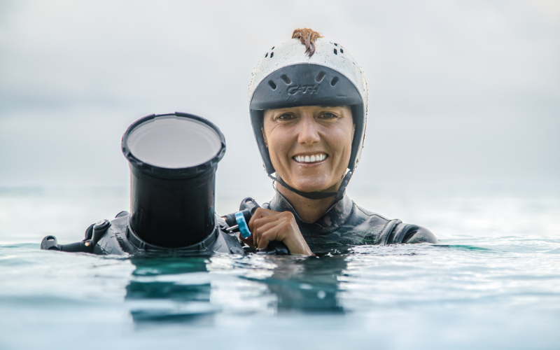 woman in wet suit in shuolder deep water holding a waterproof caerma with a large lens
