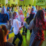 A large group of women dancing in the Stuart Community room at Ladies Dance Night. There are pink lantern decorations hanging and colored lights on the sides of the dance floor