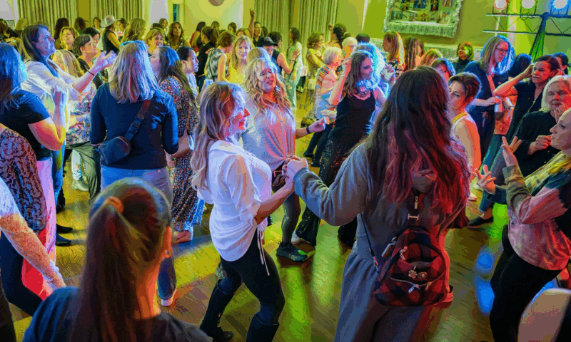 A large group of women dancing in the Stuart Community room at Ladies Dance Night. There are pink lantern decorations hanging and colored lights on the sides of the dance floor