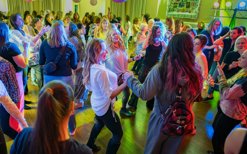 A large group of women dancing in the Stuart Community room at Ladies Dance Night. There are pink lantern decorations hanging and colored lights on the sides of the dance floor