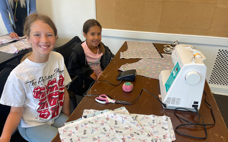 Two girls are sitting a table with a showing machine and cut fabric during the Ciao Bella Back To School sewing camp. They are smiling at the camera.
