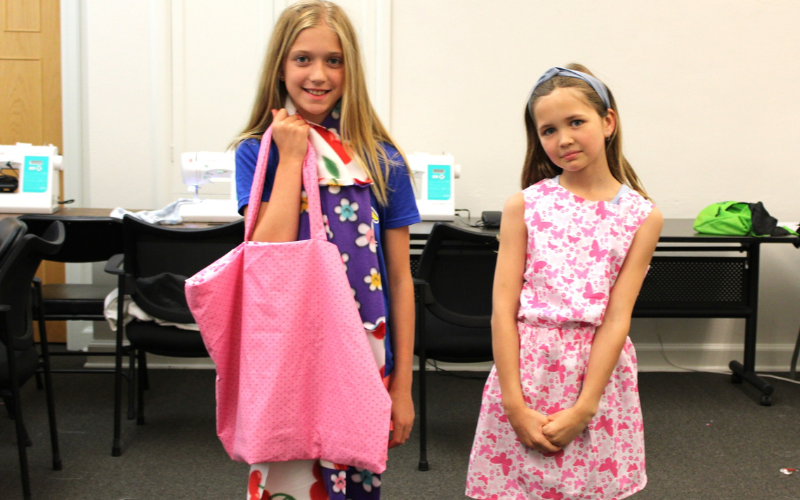 Two young girls proudly show off their creations from Ciao Bella's Sun, Sand and Stitchers class. The girl on the left made a pink tote style bag and the girl on the right is sporting a matching tank top and shorts.