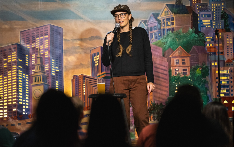 Comedian Emily Catalano is performing on stage. Behind her is a backdrop that is painted to look like a city sky line.