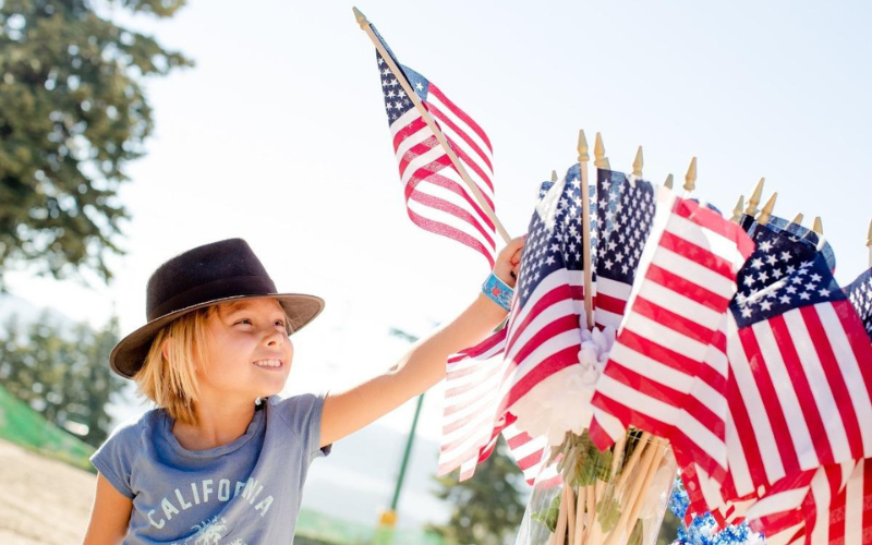 Young blond girl with a brown hat reaches for an American Flag from a bunch of flags