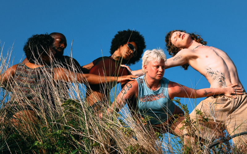Members of House of DOV are standing in tall grass with their arms outstretched, each facing different directions. There is a beautiful blue sky behind them