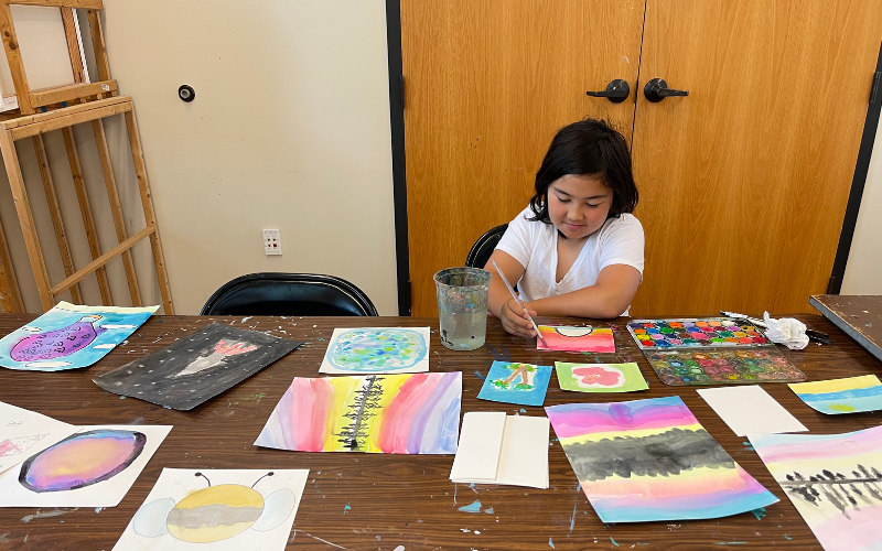 A young girl is sitting at a table, painting with watercolor paints. She is attending Art Camp with Lezlie and Carole. On the table around her are 12 other watercolor paintings.