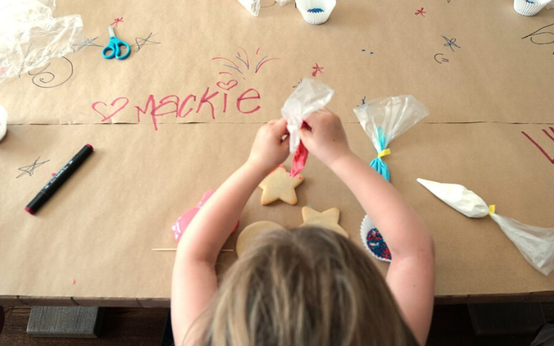 A young child is sitting at a table covered in paper. She is holding an icing bag and frosting a plain sugar cookie. The camera is above and behind her.