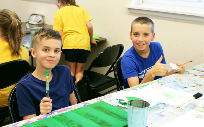 Two boys sitting at a table in Periwinkle Art Acrylic painting class. One of them has a pain project in front of him on the table and is holding a brush. They are both smiling and looking at the camera