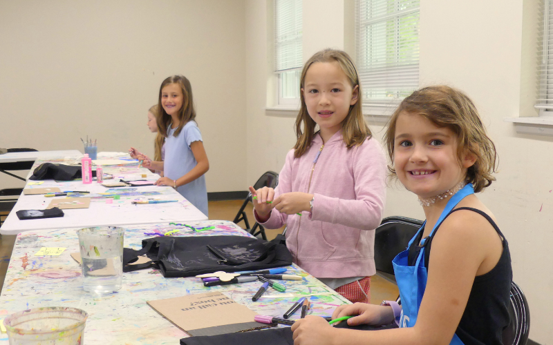 3 girls are sitting at tables working on designing tote bags in the Periwinkle: Artful Things camp. They have stopped their work to look up and smile at the camera.