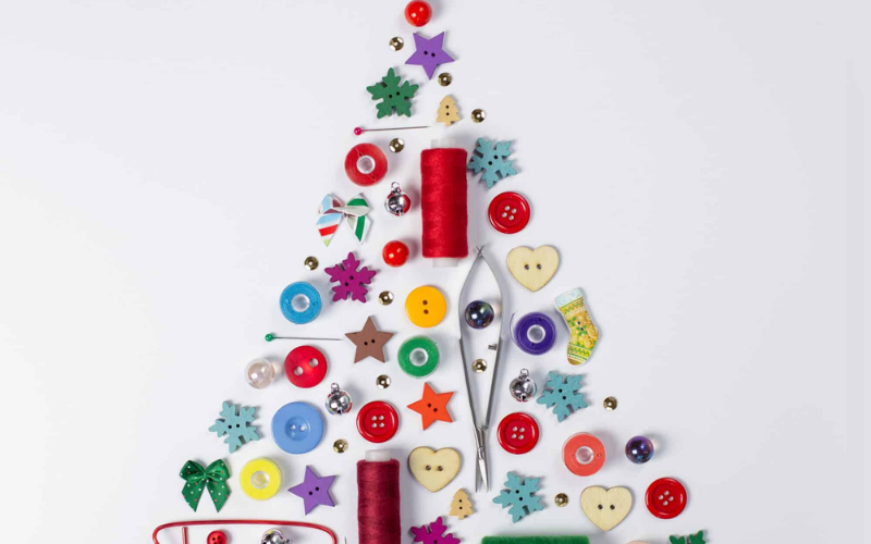 A mosaic pattern consisting of bright colored buttons, threads, beads and sewing needles arranged in the shape of a Christmas tree.