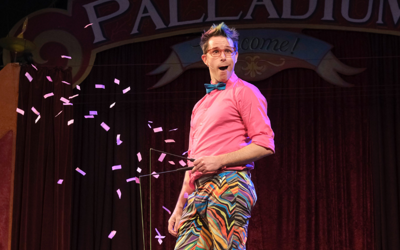 The Zaniac is performing on stage- he is dressed in bright colors, has just thrown pink confetti and is holding a diablo while looking at the audience with a surprised expression