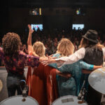 Audience gives a standing ovtion to a band that stands on stage in from of drum set after a concert in the John & Nancy Hughes Theater