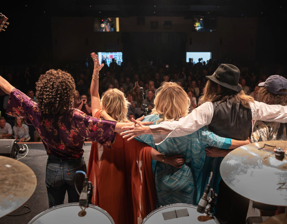 Audience gives a standing ovtion to a band that stands on stage in from of drum set after a concert in the John & Nancy Hughes Theater