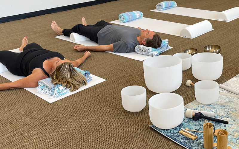 Two people laying on yoga mats on their backs, looking upward., facing away from the camera. Next to them are empty yoga mats and behind them are soundbath bowls of varying sizes.